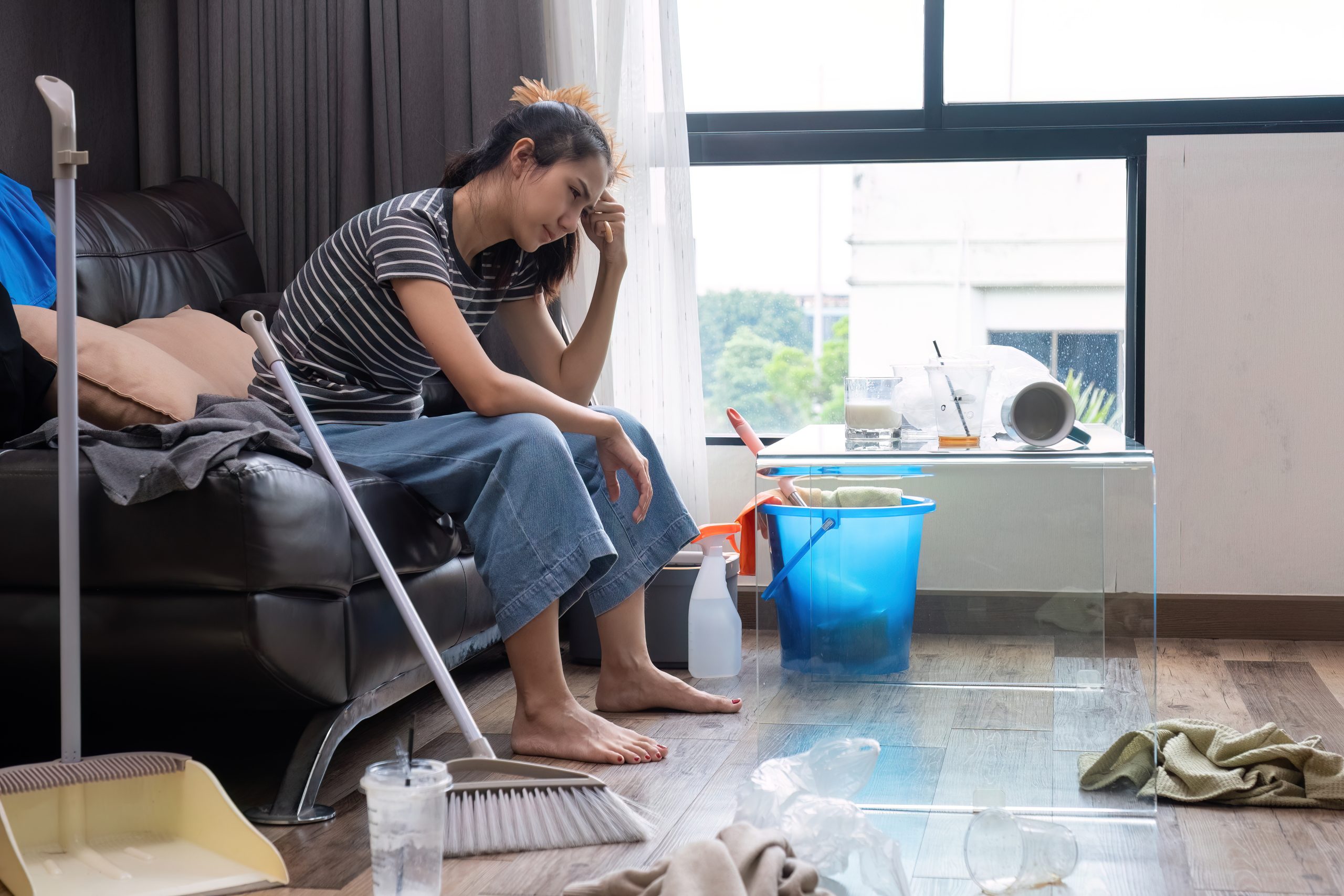 Beautiful young woman feeling tired and stressed after doing housework. She was sitting on the sofa in the living room with house cleaning supplies..